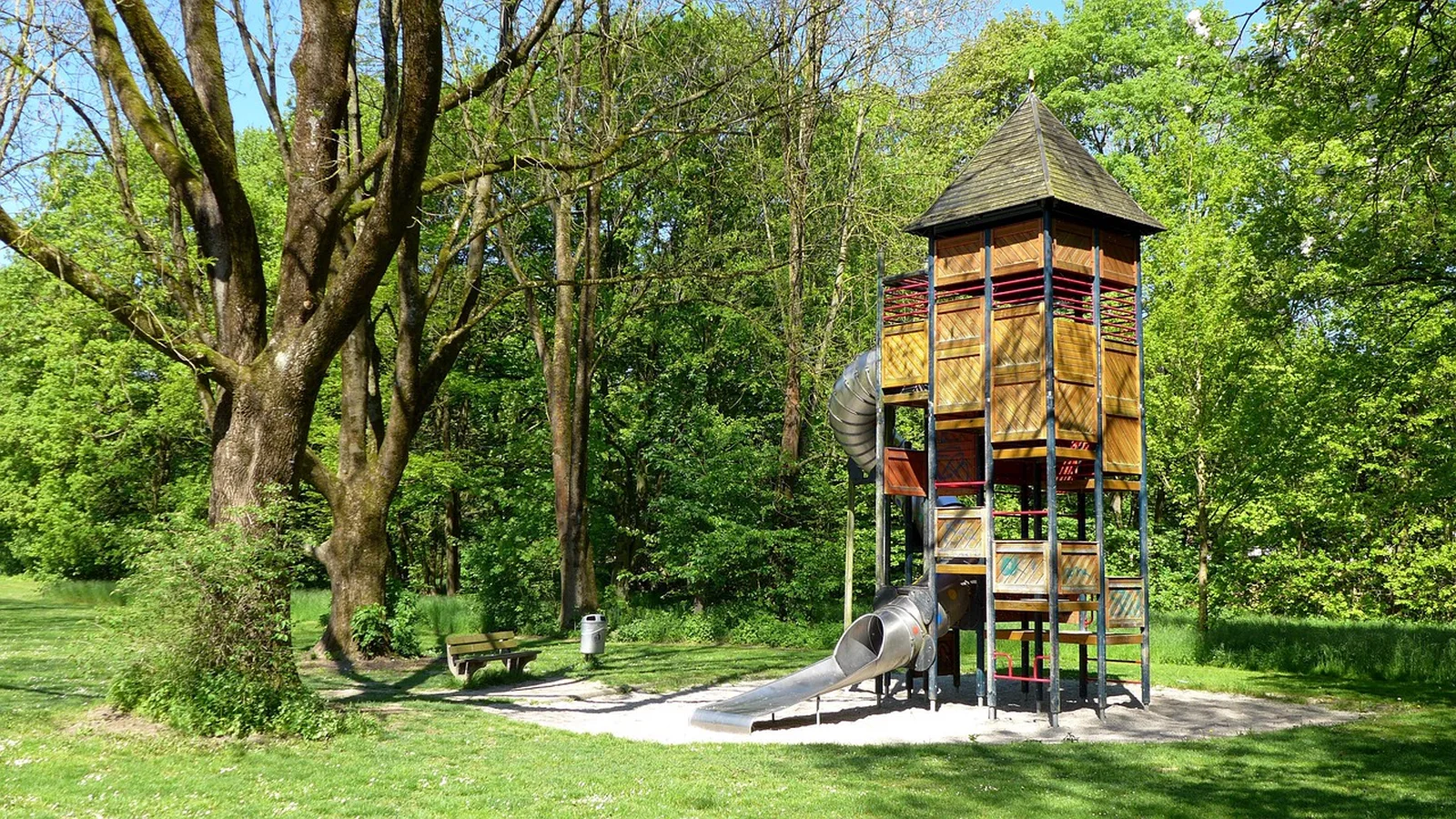 Pirate Island Sand Play: A tall wooden playground tower with a tube slide stands in a shaded grassy park.