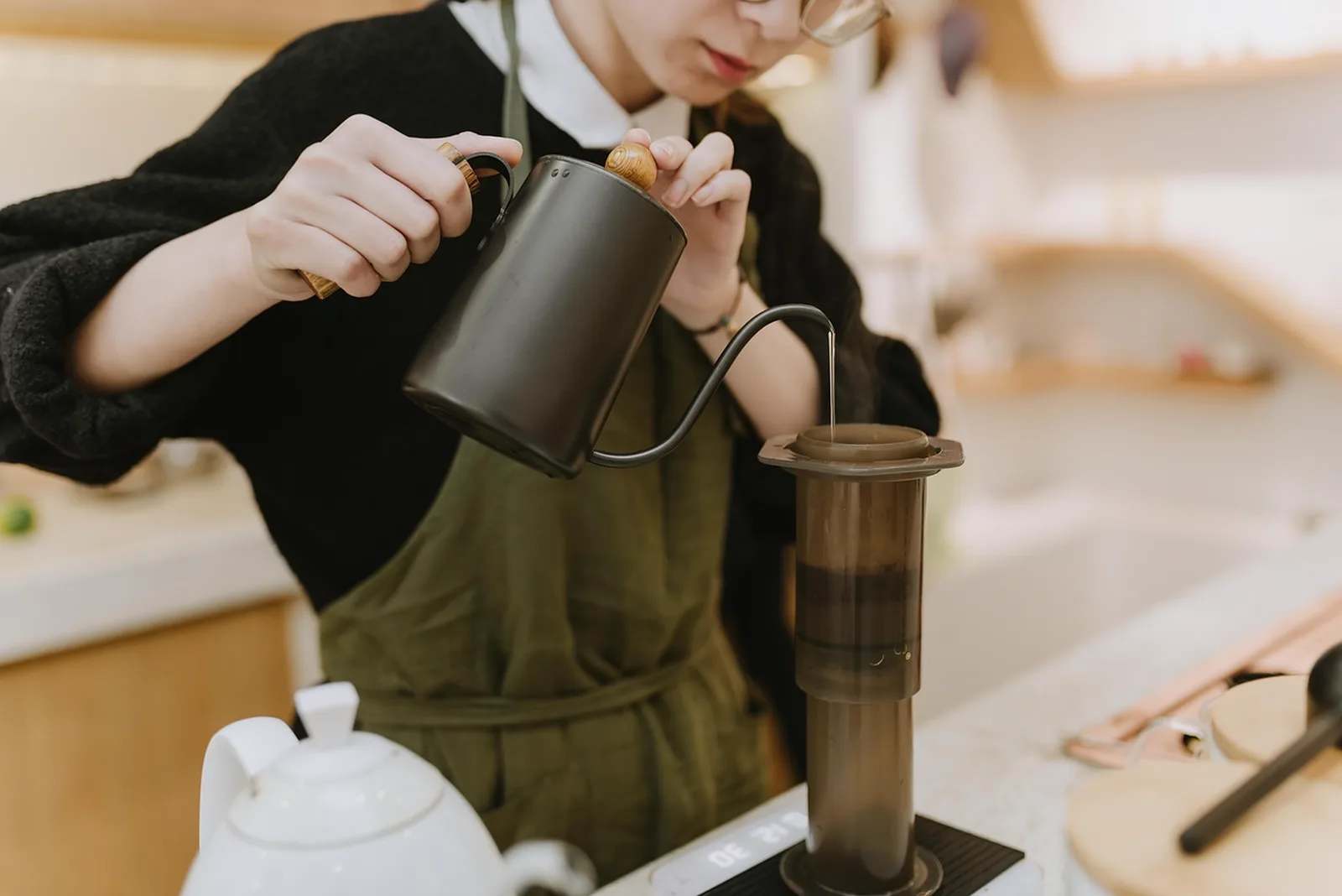 BBQ Bites: A barista pours hot water from a black gooseneck kettle into an AeroPress coffee maker on a counter.