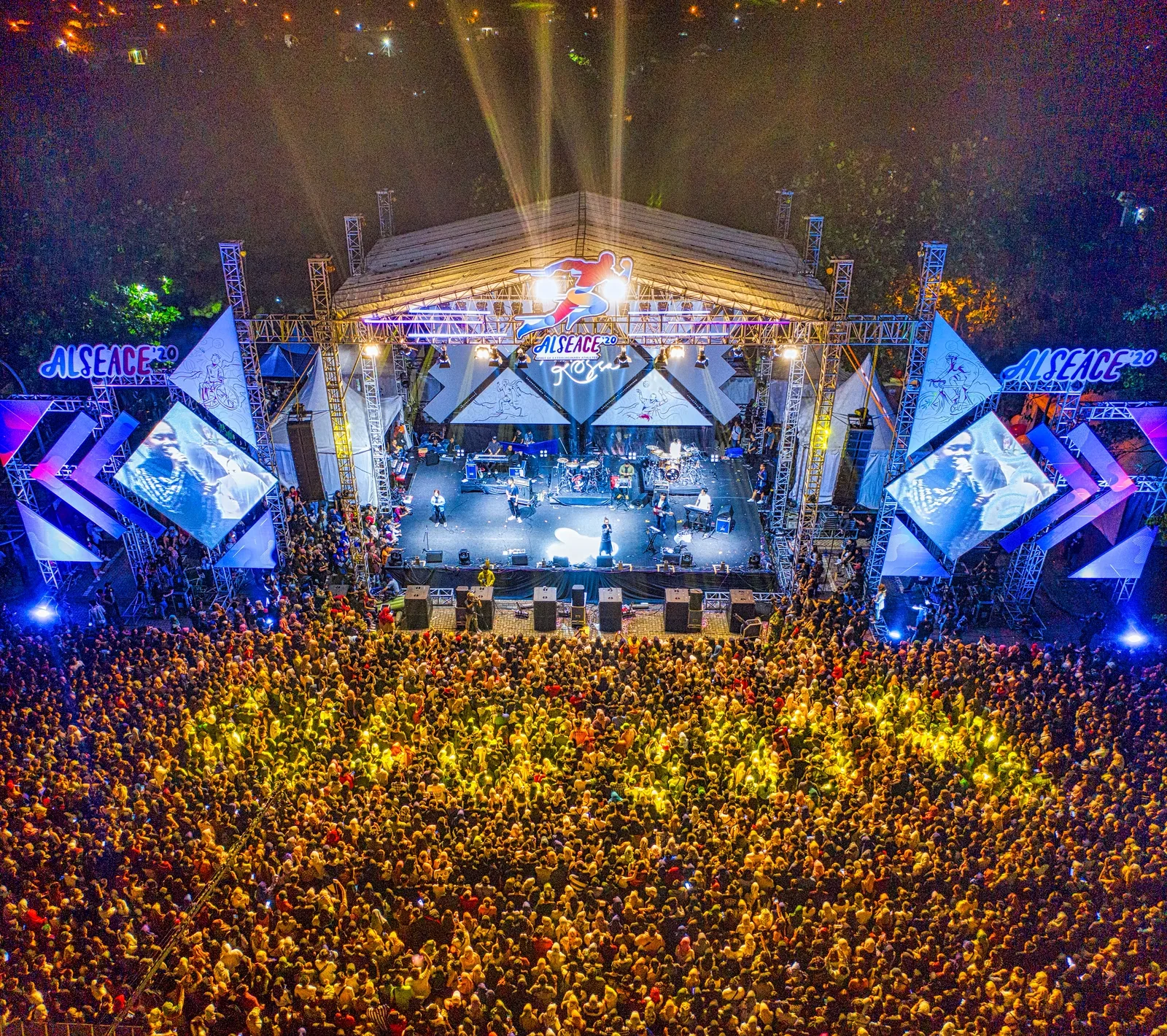 Luminous The Symphony of Us: An aerial view of an outdoor concert stage lit up at night with a large crowd gathered in front.