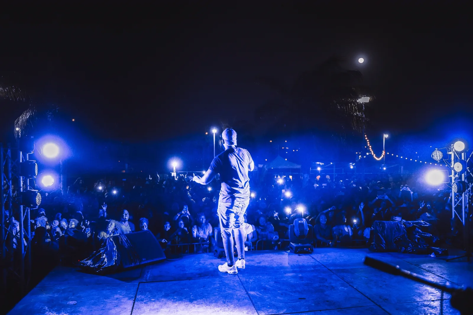 Celestial Park Nighttime Atmosphere: A performer stands on an outdoor stage illuminated by blue lights facing a crowd at night.