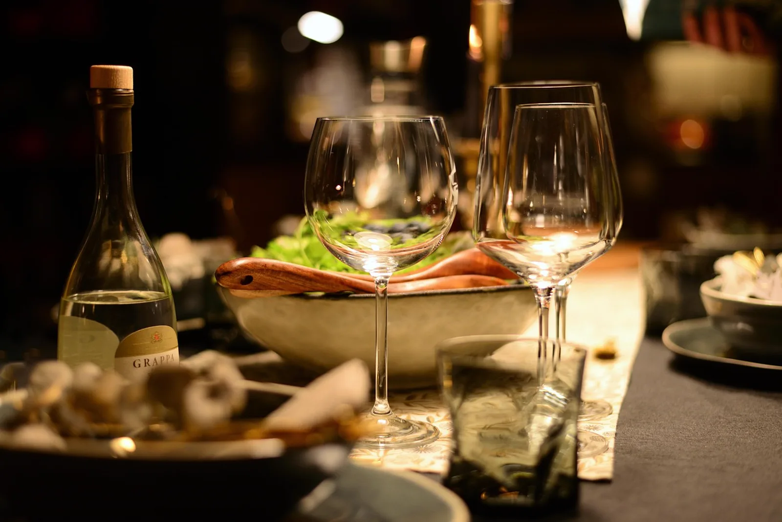 Tiffins Restaurant: Wine glasses, a salad bowl, and a bottle of grappa arranged on an elegantly set dining table in soft lighting.