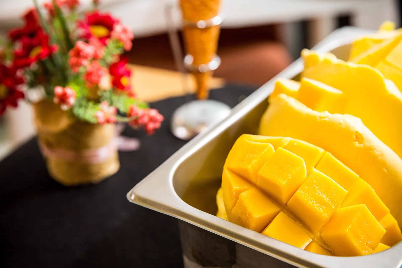 Voodoo Doughnut: A tray of yellow mango gelato with diced mango on top sitting on a counter near a blurred ice cream cone holder.