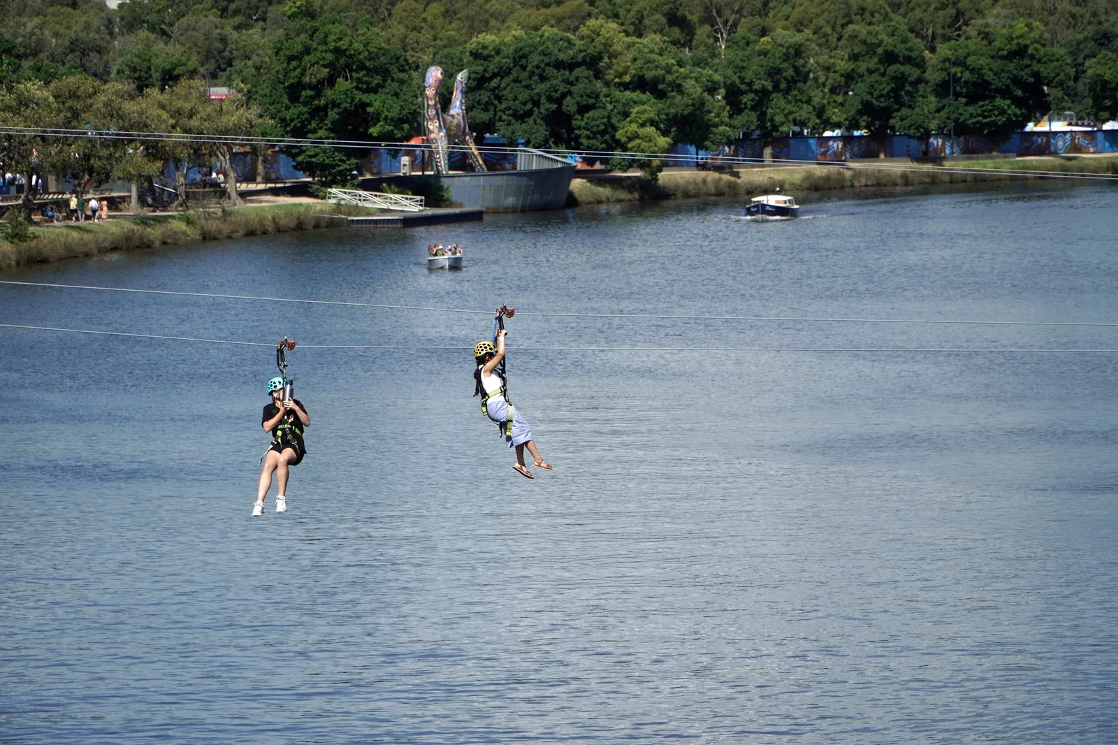Gator Gauntlet Zip Line: Two people wearing helmets zip line high above a wide river near a wooded shore.