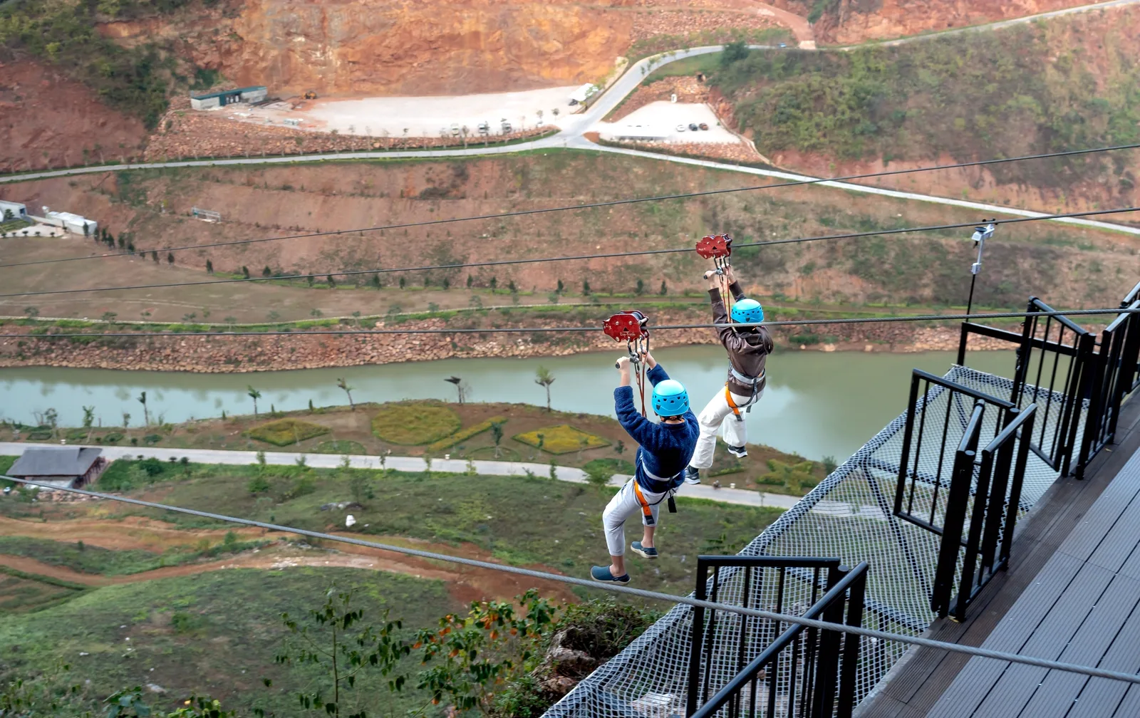 Jungala: Two people wearing blue safety helmets ride zip lines over a wide river valley.